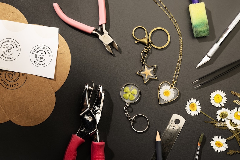 May include: An overhead shot of craft supplies and handmade jewelry. Items include a star-shaped keychain, a heart-shaped daisy pendant, a four-leaf clover keychain, and tools like pliers and a hole punch. The image also shows the text "Cotswold Crafts by Emma" on a sticker.
