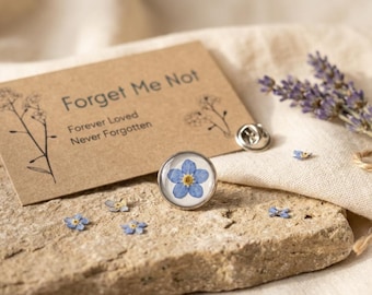 May include: A circular lapel pin featuring a pressed blue forget-me-not flower with a yellow center, displayed next to a tan card printed with the text Forget Me Not, Forever Loved, Never Forgotten, and small line drawings of flowers.