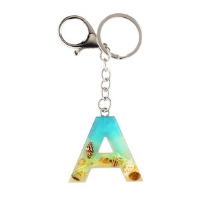 May include: A silver keyring with a blue and yellow resin letter A charm. The charm has seashells and sand embedded in it.