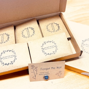 May include: A cardboard box filled with several small, square packages. Each package has a floral wreath design with the words "Forget Me Not" printed inside. A small card with the same text and a blue flower is in front.