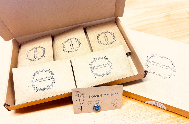 May include: A cardboard box filled with several small, square packages. Each package has a floral wreath design with the words "Forget Me Not" printed inside. A small card with the same text and a blue flower is in front.