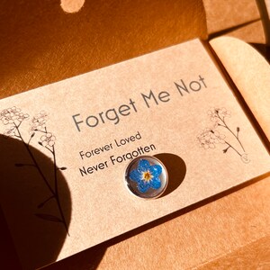 May include: A Forget Me Not pin with a real blue and yellow flower encased in clear resin. The pin is on a card with the words "Forget Me Not, Forever Loved, Never Forgotten."