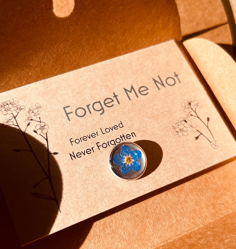 May include: A Forget Me Not pin with a real blue and yellow flower encased in clear resin. The pin is on a card with the words "Forget Me Not, Forever Loved, Never Forgotten."
