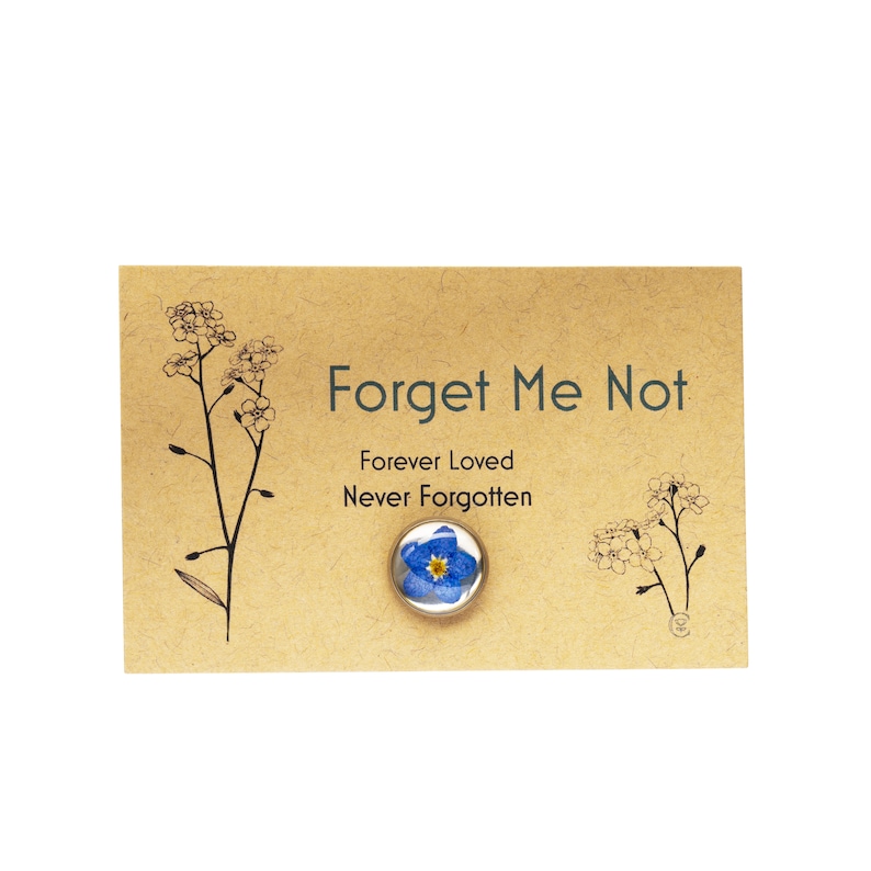 May include: A Forget Me Not pin with a blue and yellow flower design, set on a tan card. The card has the words "Forget Me Not" and "Forever Loved Never Forgotten" printed on it, along with floral illustrations.