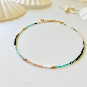 May include: A beaded necklace featuring a sequence of small round beads in black, white, turquoise, and pale pink, accented with small metallic gold beads. The necklace includes a gold-toned metal chain extender and a lobster claw clasp closure.