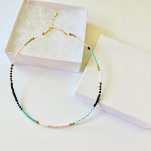 May include: A delicate beaded necklace featuring a sequence of small round beads in black, white, turquoise, and soft peach, accented with tiny gold-toned metallic spacers. The necklace includes a gold-toned metal lobster clasp and an adjustable chain.