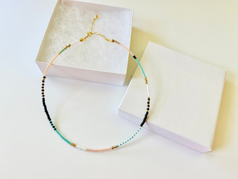 May include: A delicate beaded necklace featuring a sequence of small round beads in black, white, turquoise, and soft peach, accented with tiny gold-toned metallic spacers. The necklace includes a gold-toned metal lobster clasp and an adjustable chain.