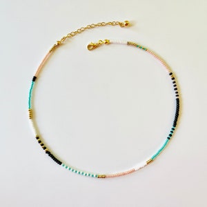 May include: A beaded necklace featuring a sequence of small, round beads in black, white, turquoise, and soft pink, accented with metallic gold-toned beads. The necklace includes a gold-toned lobster claw clasp and an adjustable chain extender.
