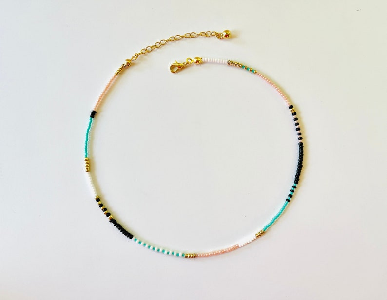 May include: A beaded necklace featuring a sequence of small, round beads in black, white, turquoise, and soft pink, accented with metallic gold-toned beads. The necklace includes a gold-toned lobster claw clasp and an adjustable chain extender.