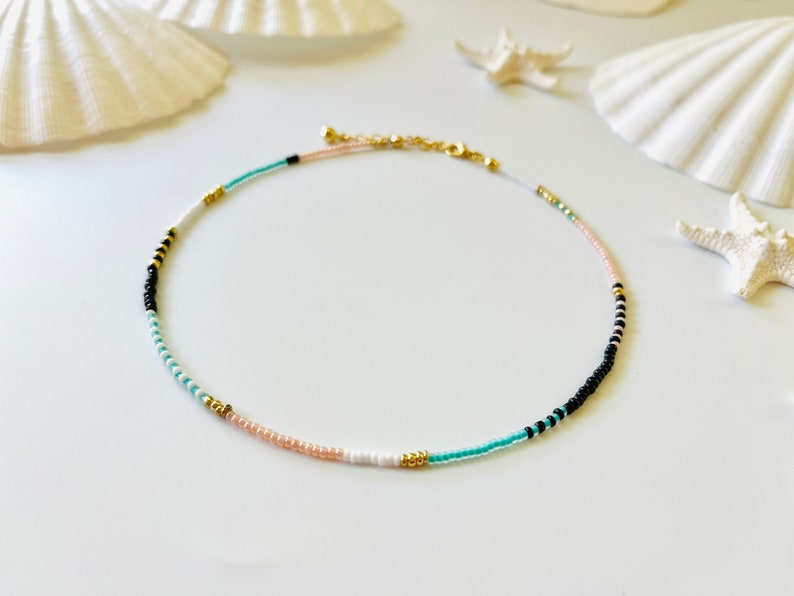 May include: A beaded necklace featuring a sequence of small round beads in black, white, turquoise, and pale pink, accented with small metallic gold beads. The necklace includes a gold-toned metal chain extender and a lobster claw clasp closure.