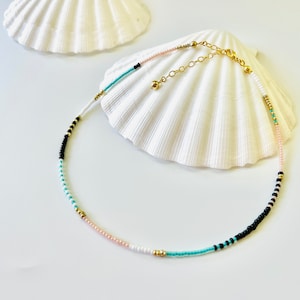 May include: A beaded necklace featuring a sequence of small round beads in black, white, light pink, and turquoise, accented with metallic gold beads. The necklace includes a gold-toned metal chain extender with a small gold bead at the end.