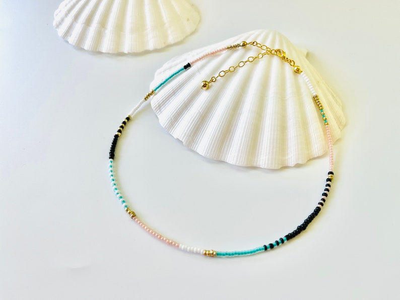 May include: A beaded necklace featuring a sequence of small round beads in black, white, light pink, and turquoise, accented with metallic gold beads. The necklace includes a gold-toned metal chain extender with a small gold bead at the end.