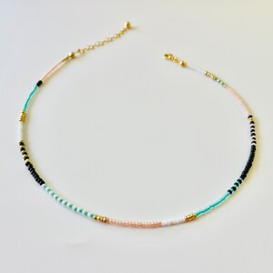 May include: A delicate beaded necklace featuring a sequence of small, round beads in shades of black, white, turquoise, and pale pink, accented with small gold-toned metallic beads. The necklace includes a gold-toned chain extender and a lobster clasp.