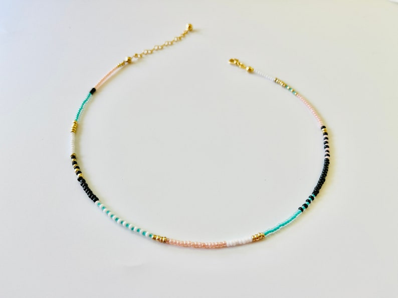 May include: A delicate beaded necklace featuring a sequence of small, round beads in shades of black, white, turquoise, and pale pink, accented with small gold-toned metallic beads. The necklace includes a gold-toned chain extender and a lobster clasp.