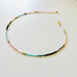 May include: A delicate beaded necklace featuring a sequence of small, colorful seed beads in shades of turquoise, black, white, peach, and metallic gold. The necklace includes a gold-toned metal chain extender and a small lobster claw clasp closure.