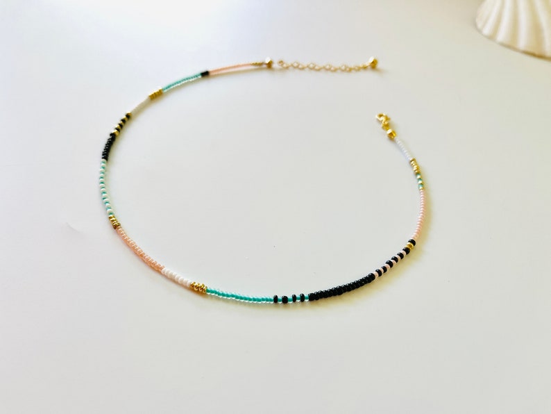 May include: A delicate beaded necklace featuring a sequence of small, colorful seed beads in shades of turquoise, black, white, peach, and metallic gold. The necklace includes a gold-toned metal chain extender and a small lobster claw clasp closure.