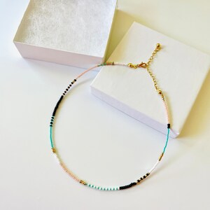 May include: A delicate beaded necklace featuring a pattern of small white, black, turquoise, light pink, and metallic gold beads. The necklace includes a gold-toned metal lobster clasp and an adjustable chain extender, presented with a white box.