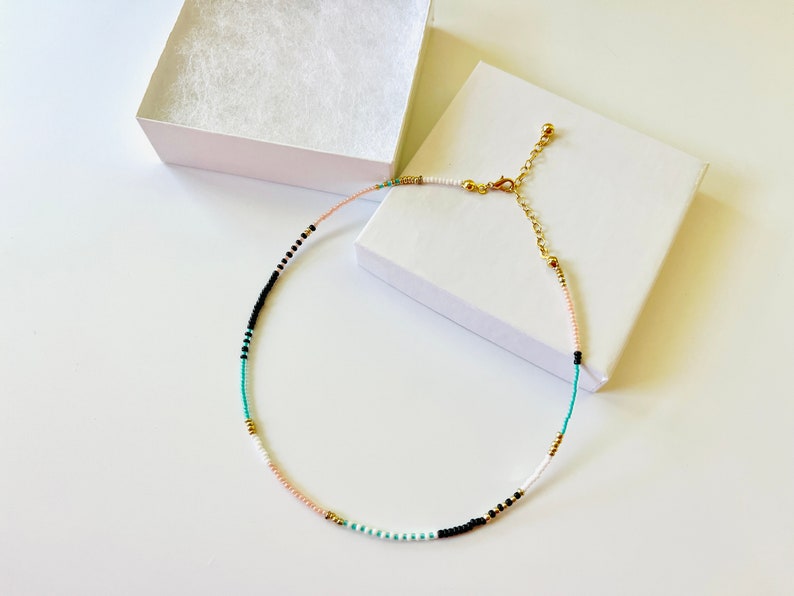 May include: A delicate beaded necklace featuring a pattern of small white, black, turquoise, light pink, and metallic gold beads. The necklace includes a gold-toned metal lobster clasp and an adjustable chain extender, presented with a white box.