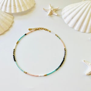May include: A beaded necklace featuring a repeating pattern of small white, black, turquoise, and pale pink seed beads interspersed with tiny metallic gold spacer beads. The necklace includes a gold-toned metal lobster clasp and an adjustable chain.