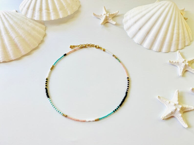 May include: A beaded necklace featuring a repeating pattern of small white, black, turquoise, and pale pink seed beads interspersed with tiny metallic gold spacer beads. The necklace includes a gold-toned metal lobster clasp and an adjustable chain.