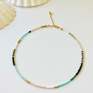 May include: A delicate beaded necklace featuring a sequence of small round beads in shades of black, white, turquoise, and soft pink, accented with metallic gold-toned beads. The necklace includes a gold-toned metal chain extender and a lobster clasp.