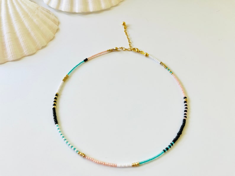 May include: A delicate beaded necklace featuring a sequence of small round beads in shades of black, white, turquoise, and soft pink, accented with metallic gold-toned beads. The necklace includes a gold-toned metal chain extender and a lobster clasp.