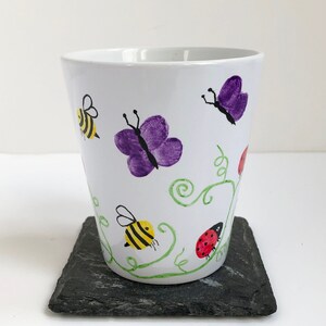 May include: White ceramic mug with hand-painted purple butterflies, yellow and black bees, and red ladybugs on a green vine design.