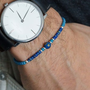 May include: A blue and turquoise beaded bracelet with a gold charm on a person's wrist. The bracelet is worn with a silver watch with a white face and a black leather band.