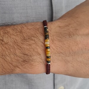 May include: A beaded bracelet featuring a series of small, cylindrical beads in shades of brown, yellow, and black. The bracelet is strung on a dark red cord and secured with a woven knot. The bracelet is shown on a person's wrist.