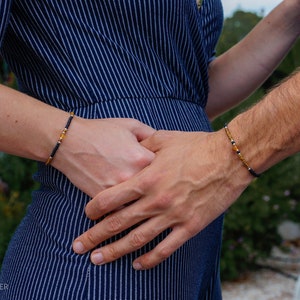 Puede incluir: Dos pulseras de cuentas con cuentas negras y doradas se llevan en las muñecas. Las pulseras tienen un cordón azul oscuro. La imagen muestra a dos personas tomadas de la mano, con las pulseras visibles en sus muñecas. El fondo está borroso.