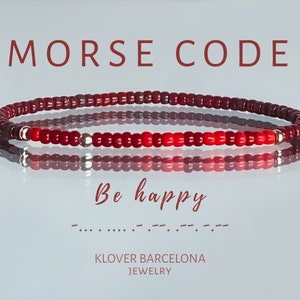 Puede incluir: Pulsera de cuentas rojas con cuentas plateadas, que deletrea "Be happy" en código Morse. La pulsera está sobre una superficie blanca reflectante. "MORSE CODE" y "KLOVER BARCELONA JEWELRY" están escritos en rojo arriba y abajo de la pulsera.
