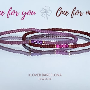 Może przedstawiać: Trzy bransoletki z koralików w kolorze fioletowym i bordowym z złotymi akcentami i małymi fioletowymi kamieniami szlachetnymi. Bransoletki są ułożone jedna na drugiej, a nad nimi widoczny jest tekst "One for you One for me".