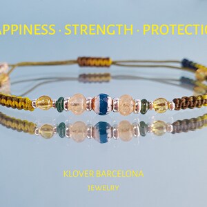 Op de afbeelding: Een groene, gele en bruine armband van touw met een blauwe en witte agaatkraal, heldere kwarts kralen en gouden kralen. De armband heeft de woorden "Happiness, Strength, Protection" erboven geschreven.