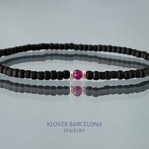 Può includere: Un braccialetto di perline nere con una gemma di rubino centrale. Il braccialetto è composto da piccole perline nere quadrate e un rubino rosso vivo. Il braccialetto è presentato su una superficie riflettente. Il marchio "KLOVER BARCELONA JEWELRY" è visibile.