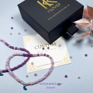 Puede incluir: Dos pulseras de cuentas moradas y lavanda con dijes plateados en forma de corazón. Las pulseras están sobre una superficie blanca con una tarjeta que dice "CUSTOM" y "KLOVER BARCELONA JEWELRY".