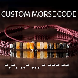 May include: A red braided bracelet with a row of colorful beads and a silver charm. The bracelet is reflected in a dark surface. The text "CUSTOM MORSE CODE" is displayed above the bracelet. The morse code ".. -.. .. -.. - -.. - - - - -" is displayed below the bracelet.