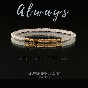 May include: White beaded bracelet with a gold bar charm. The bracelet is on a black background and the word "Always" is written above it in a cursive font. The text "KLOVER BARCELONA JEWELRY" is written below the bracelet.