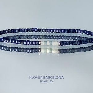 May include: A blue and white beaded bracelet with a silver accent. The bracelet is made of small, round beads and has a simple, elegant design. KLOVER BARCELONA JEWELRY is printed below the bracelet.