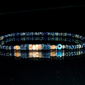 Puede incluir: Una pulsera de cuentas con una mezcla de cuentas azules, beige y plateadas. La pulsera incluye una sección central de cuentas rectangulares y un pequeño dije de ojo azul. La pulsera se muestra sobre una superficie negra reflectante.