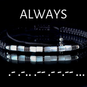 Pode incluir: Um bracelete de cordão preto com contas brancas e prateadas. O bracelete tem a palavra "ALWAYS" escrita acima e uma linha de pontos abaixo. O bracelete é refletido em uma superfície preta.