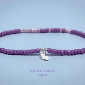 Puede incluir: Un brazalete de cuentas moradas y rosas con un dije de corazón plateado. El brazalete está hecho con cuentas pequeñas y redondas y tiene un aspecto delicado y femenino.