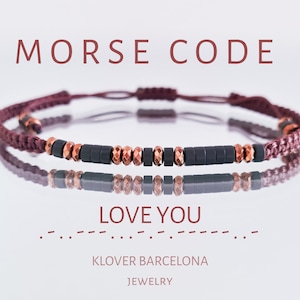 Pode incluir: Um bracelete de cordão marrom com contas pretas e cobre que soletram "LOVE YOU" em código Morse. O bracelete está sobre um fundo branco com o texto "MORSE CODE" e "KLOVER BARCELONA JEWELRY" abaixo.