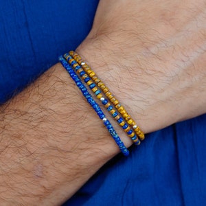 Puede incluir: Tres pulseras de cuentas en tonos azules y dorados se llevan en la muñeca. Las pulseras están hechas de pequeñas cuentas redondas. El fondo es de tela azul lisa.