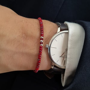 Peut inclure: Un bracelet rouge en perles avec des accents argentés porté sur un poignet avec un cadran de montre blanc et un bracelet en cuir marron.