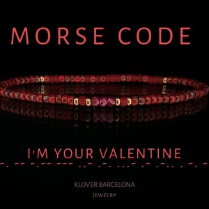 Puede incluir: Un brazalete de cuentas rojas con detalles dorados y un mensaje en código Morse que dice "Soy tu Valentín". El brazalete está sobre un fondo negro y se refleja en una superficie de espejo. El texto "KLOVER BARCELONA JEWELRY" está debajo del brazalete.