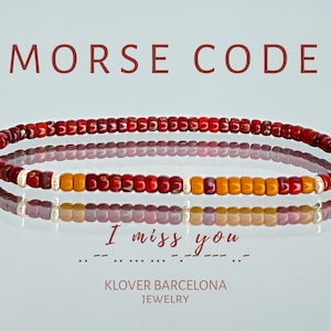 Può includere: Un braccialetto di perline rosse e arancioni con le parole "MORSE CODE" sopra. Il braccialetto mostra il codice Morse per "Mi manchi". È visibile anche il testo "KLOVER BARCELONA JEWELRY". Il braccialetto misura circa 18 cm.