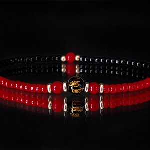 Puede incluir: Un brazalete de cuentas rojas y negras con una cuenta negra con letras doradas. El brazalete está sobre un fondo negro.