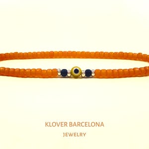 Könnte beinhalten: Orangefarbenes Armband mit Perlen und einem blauen und gelben bösen Blick-Anhänger. KLOVER BARCELONA JEWELRY.
