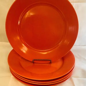 May include: A stack of six orange-colored dinner plates. The plates are round with a slightly raised rim. One plate is propped up on a decorative metal stand, showcasing the vibrant color and glossy finish. The plates are a solid color.
