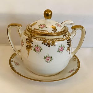 May include: A white porcelain sugar bowl with a lid, saucer, and spoon. The set is decorated with pink roses and gold accents. The handles and finial are also gold. The sugar bowl sits on a matching saucer.